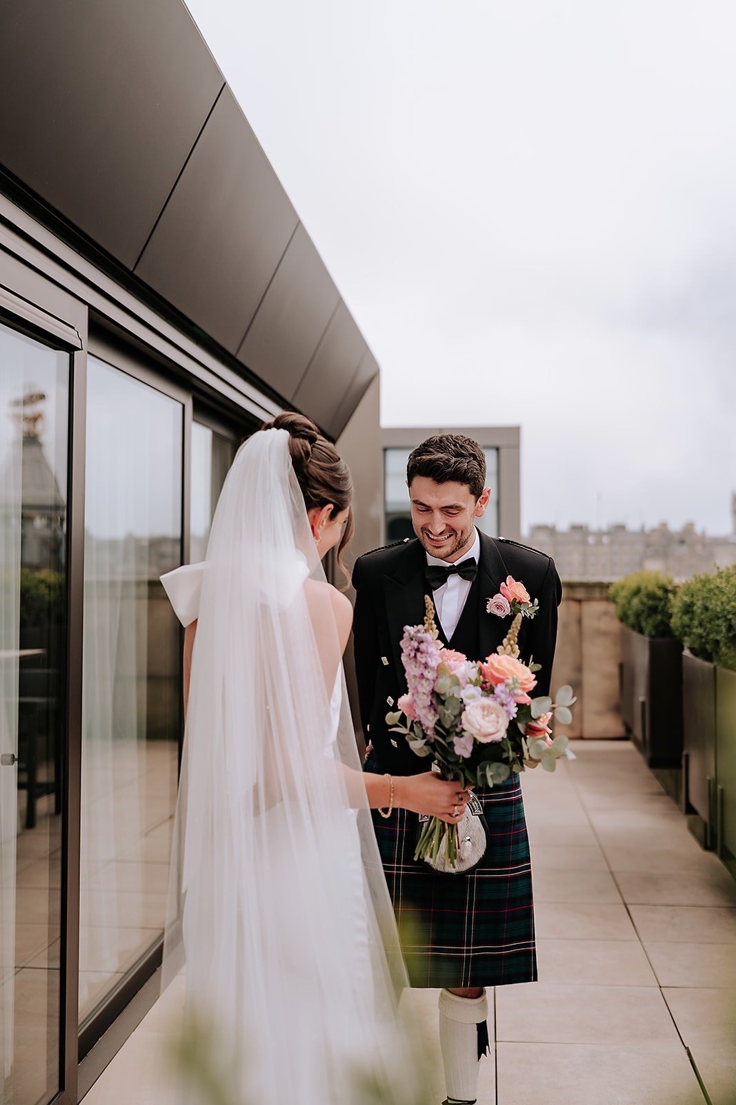 A bride and groom sharing a moment on a balcony, with the bride holding a bouquet of pink and purple flowers, the groom wearing a kilt and a tuxedo, both smiling.