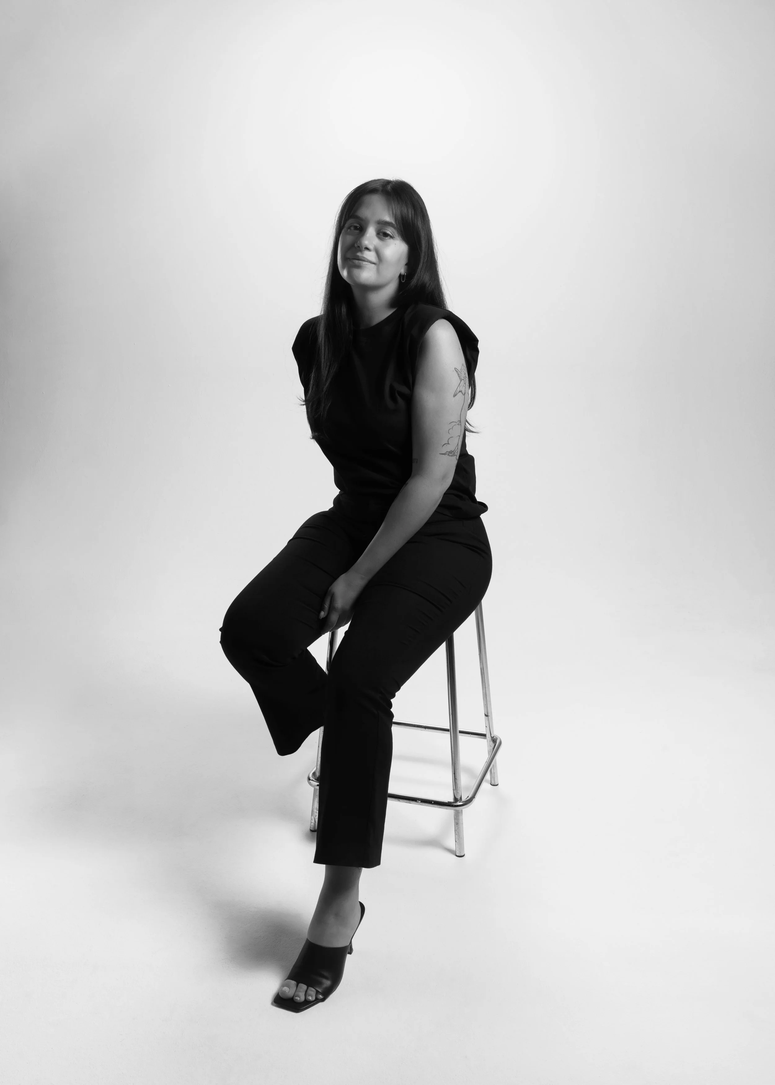 Une femme assise sur un tabouret, portant des vêtements noirs, souriant légèrement, dans un studio avec fond blanc, en noir et blanc.