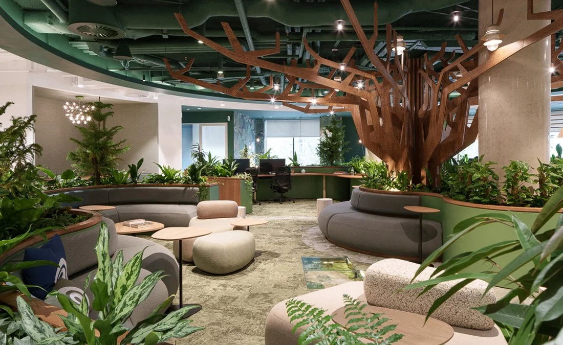 Modern office lounge with green plants, a large wooden tree sculpture on the ceiling, and curved seating areas.
