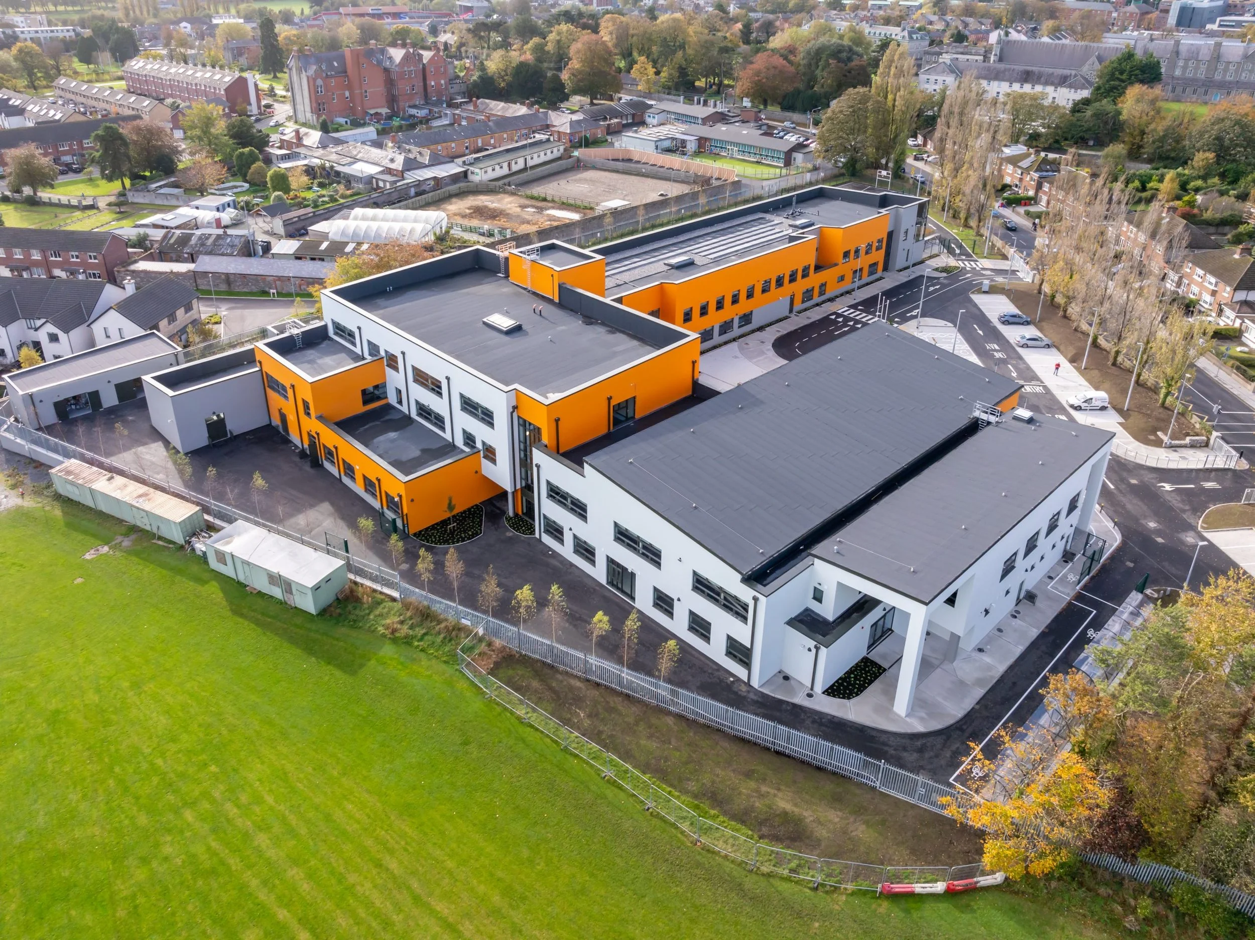 An aerial view of a modern school building with orange and white exterior, surrounded by parking lots, sidewalks, and a grassy field with trees.