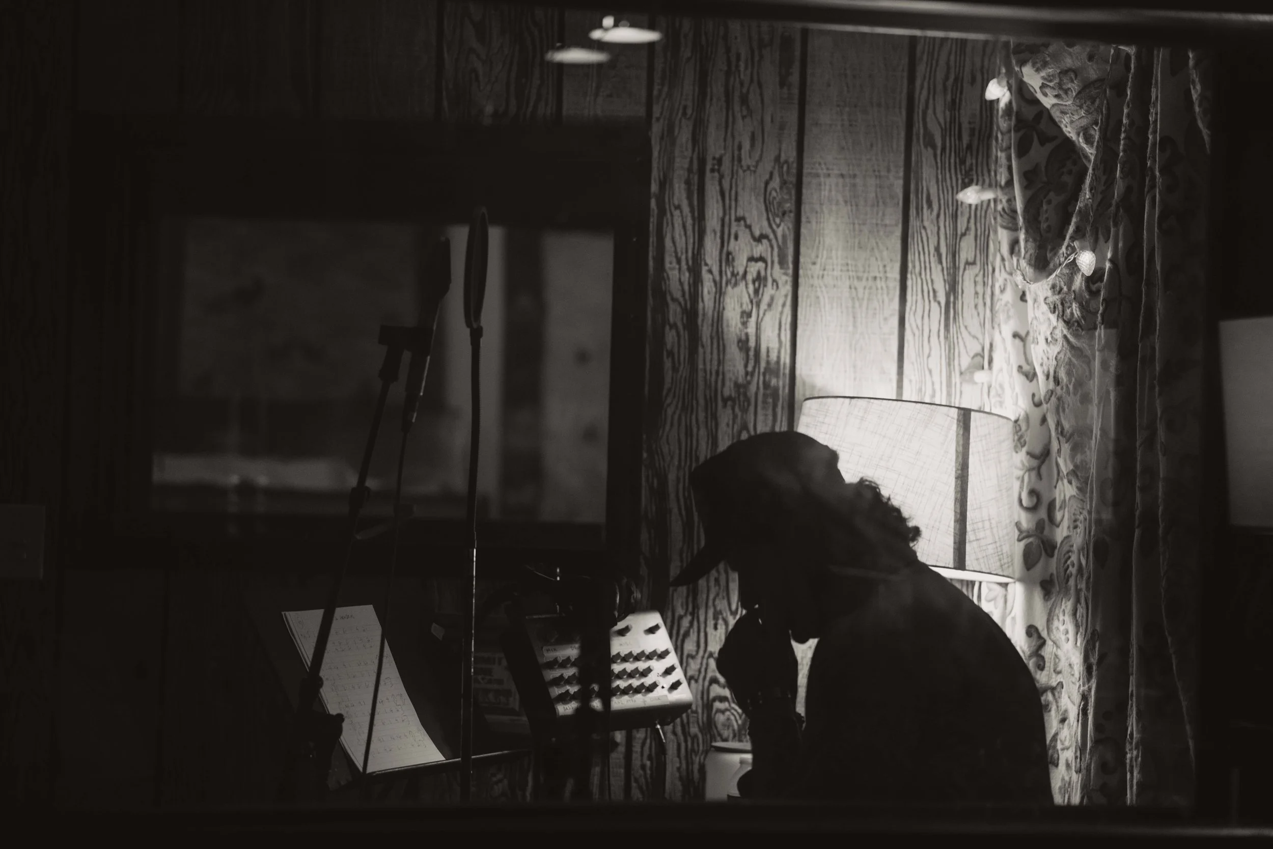 Man plays harmonica in a recording studio while making a new album, image is black and white with a lamp casting him into shadow.