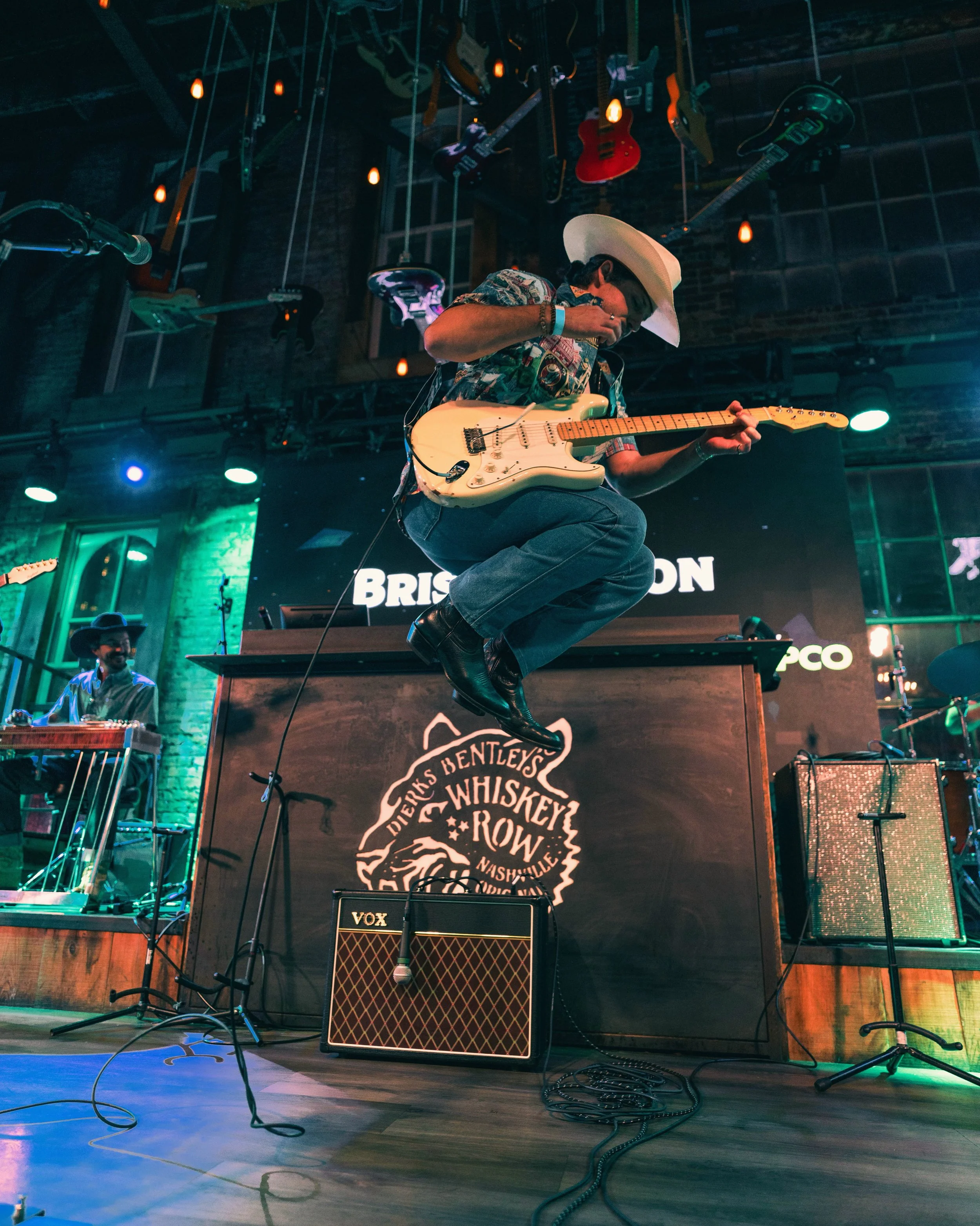 Country singer in cowboy hat jumping on stage while playing guitar with neon lights around him.