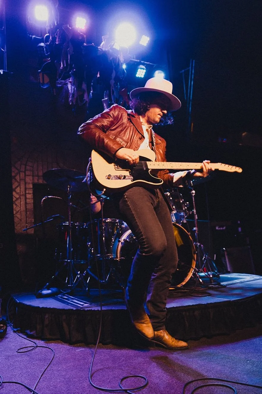 Country singer plays guitar on stage with white cowboy hat and cowboy boots on. There are blue stage lights.