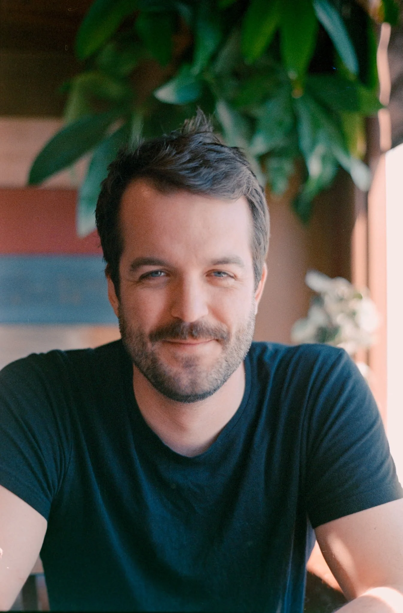 A man with dark hair and a beard sitting indoors with greenery behind him and a wooden window frame to the side, smiling at the camera.