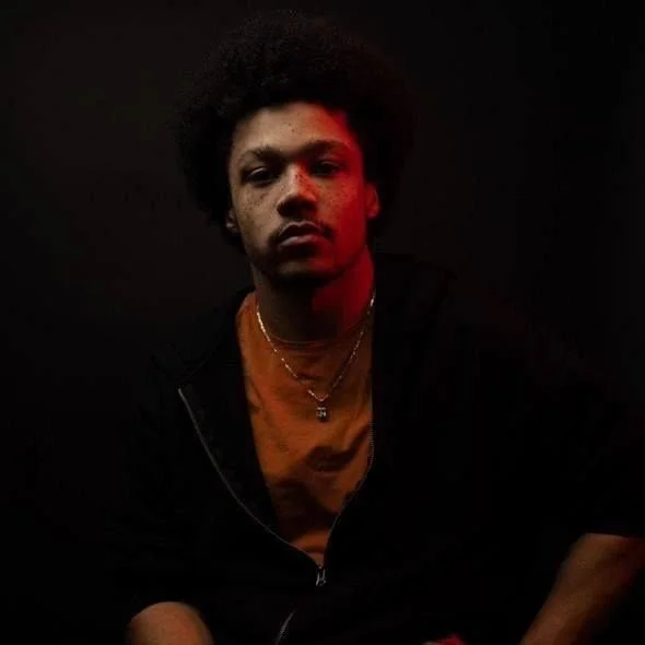 Portrait of a young man with dark curly hair, wearing a black hoodie over a brown T-shirt, against a dark background.