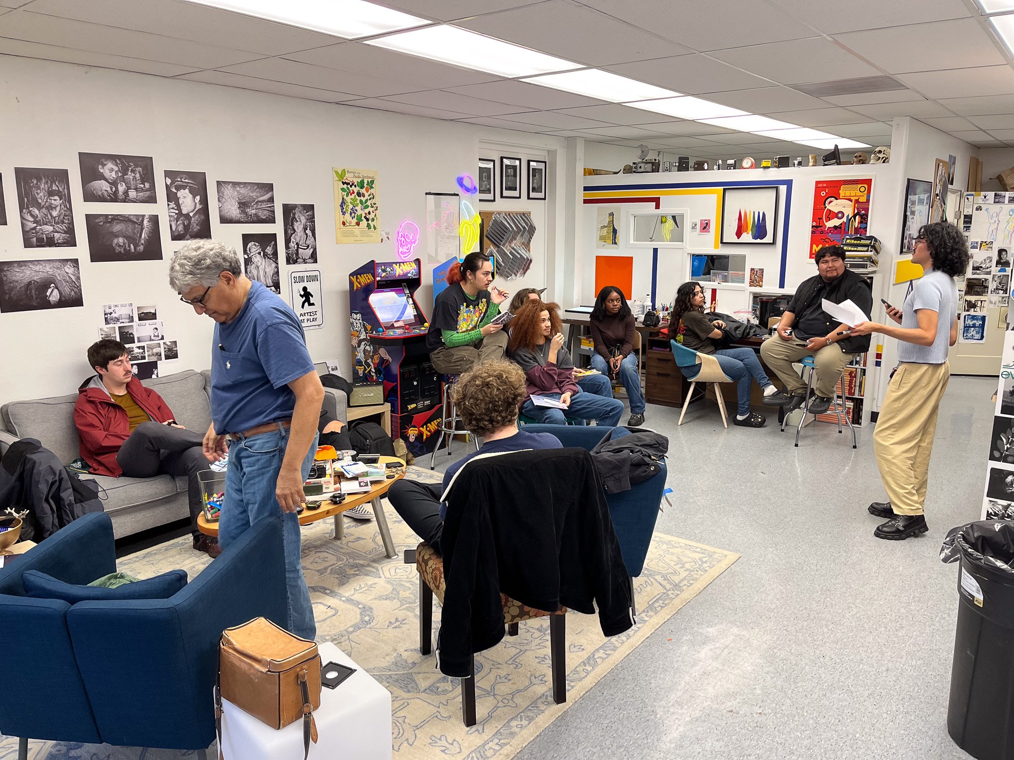 People gathered in an art studio or classroom, some sitting, some standing, listening to a woman speaking while holding papers, with art and photographs decorating the walls.