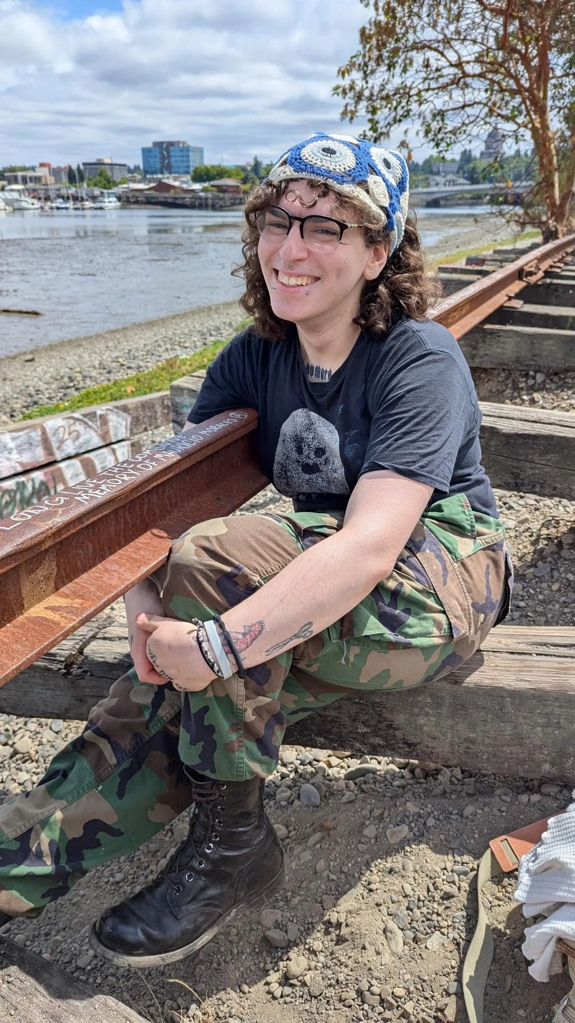 A person with curly hair, glasses, and tattoos on their arms, sitting on the ground near a railroad track by the water, smiling at the camera, wearing a colorful knit hat, black t-shirt with a face graphic, camouflage pants, and black boots, with a tree and city skyline in the background.