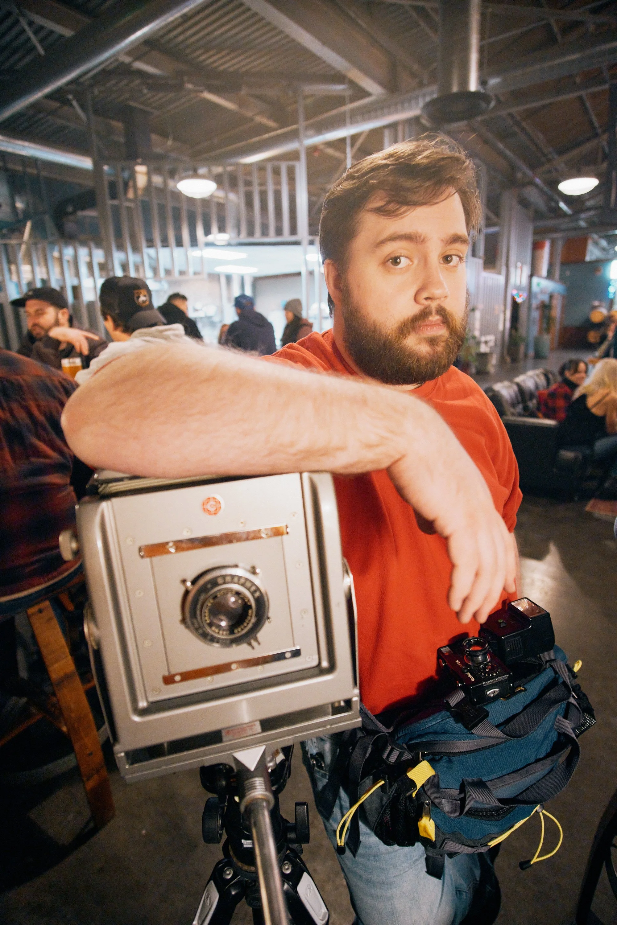 A man with a beard and red shirt sitting at a bar with a vintage camera on a tripod in front of him. The bar is busy with other patrons and has exposed ceiling pipes.