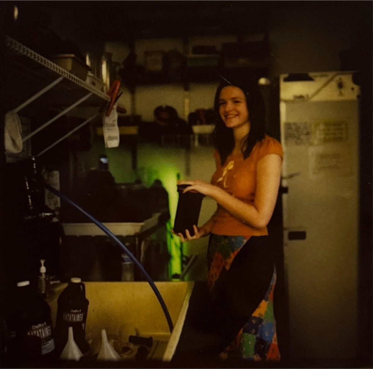 A woman with black hair, wearing an orange shirt and a colorful skirt, standing in a dark kitchen, holding a black device with green slime dripping from it.