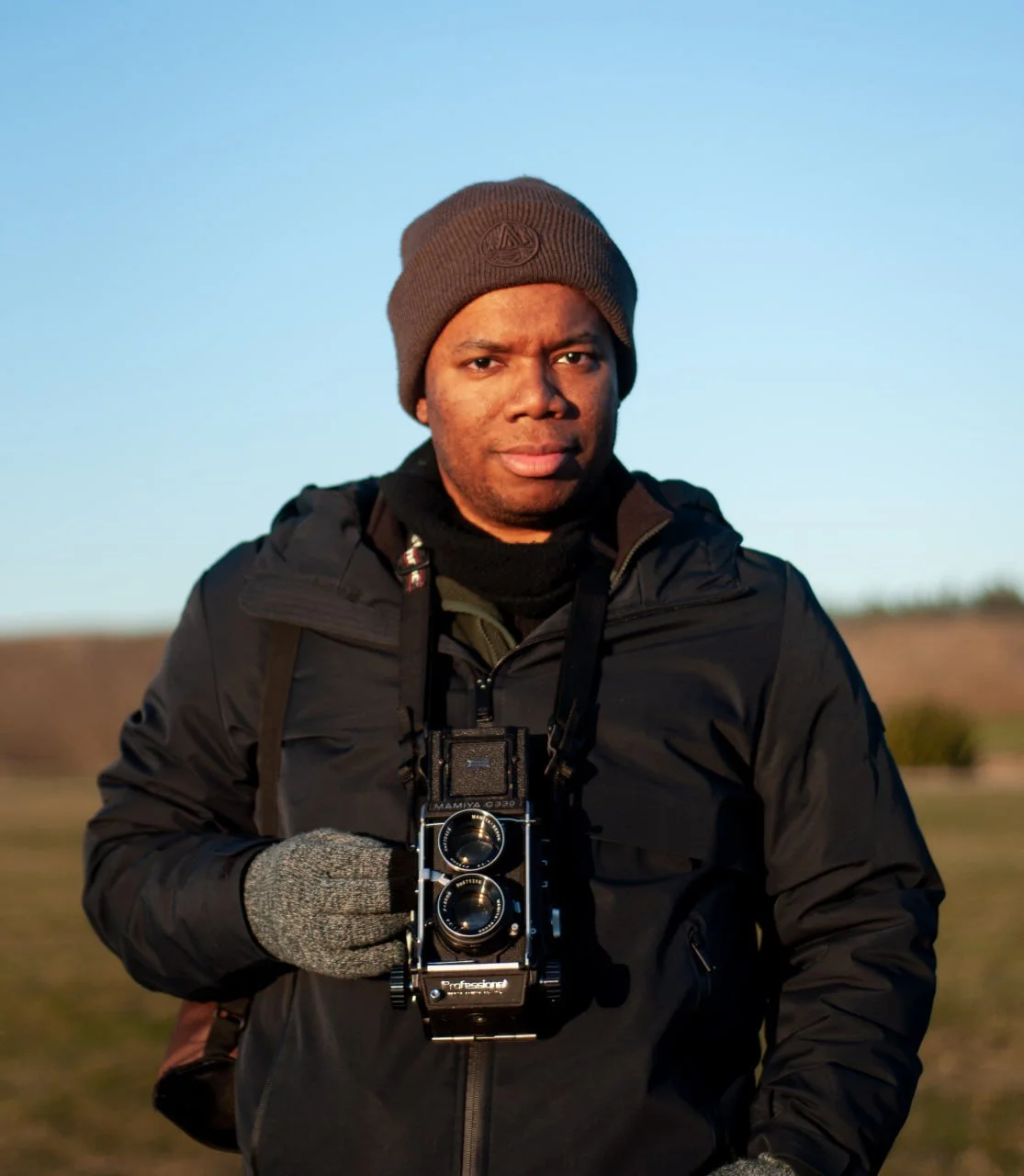 A man wearing a brown beanie, black jacket, and gray gloves, holding a vintage twin-lens reflex camera, standing outdoors with a clear blue sky in the background.