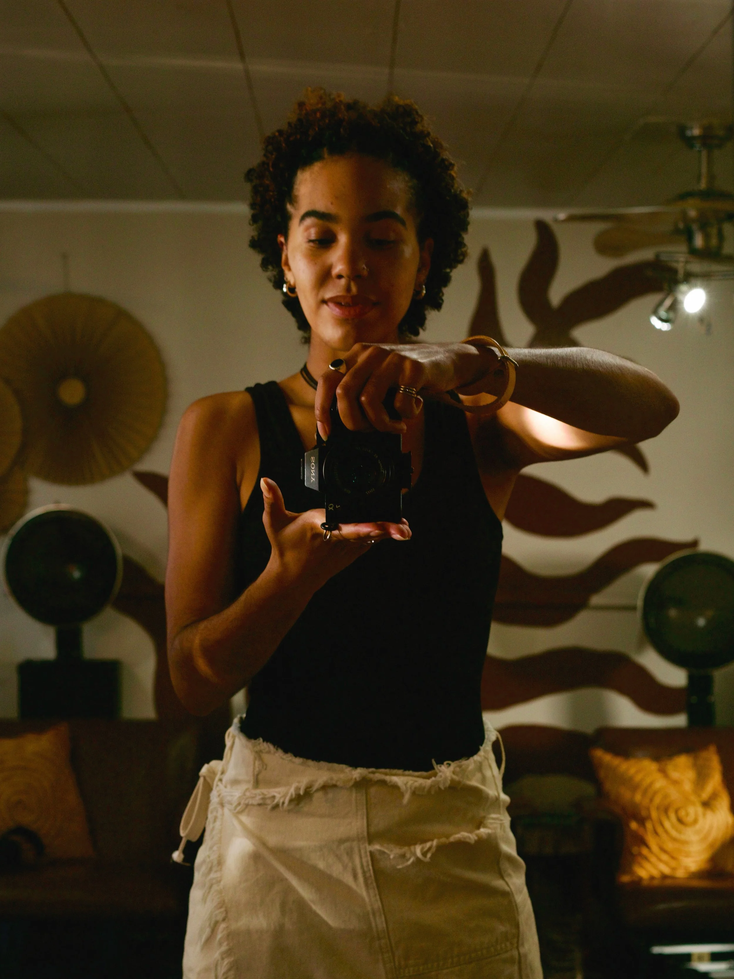 Young woman taking a mirror selfie with a camera in a dimly lit room decorated with wall art and speakers.