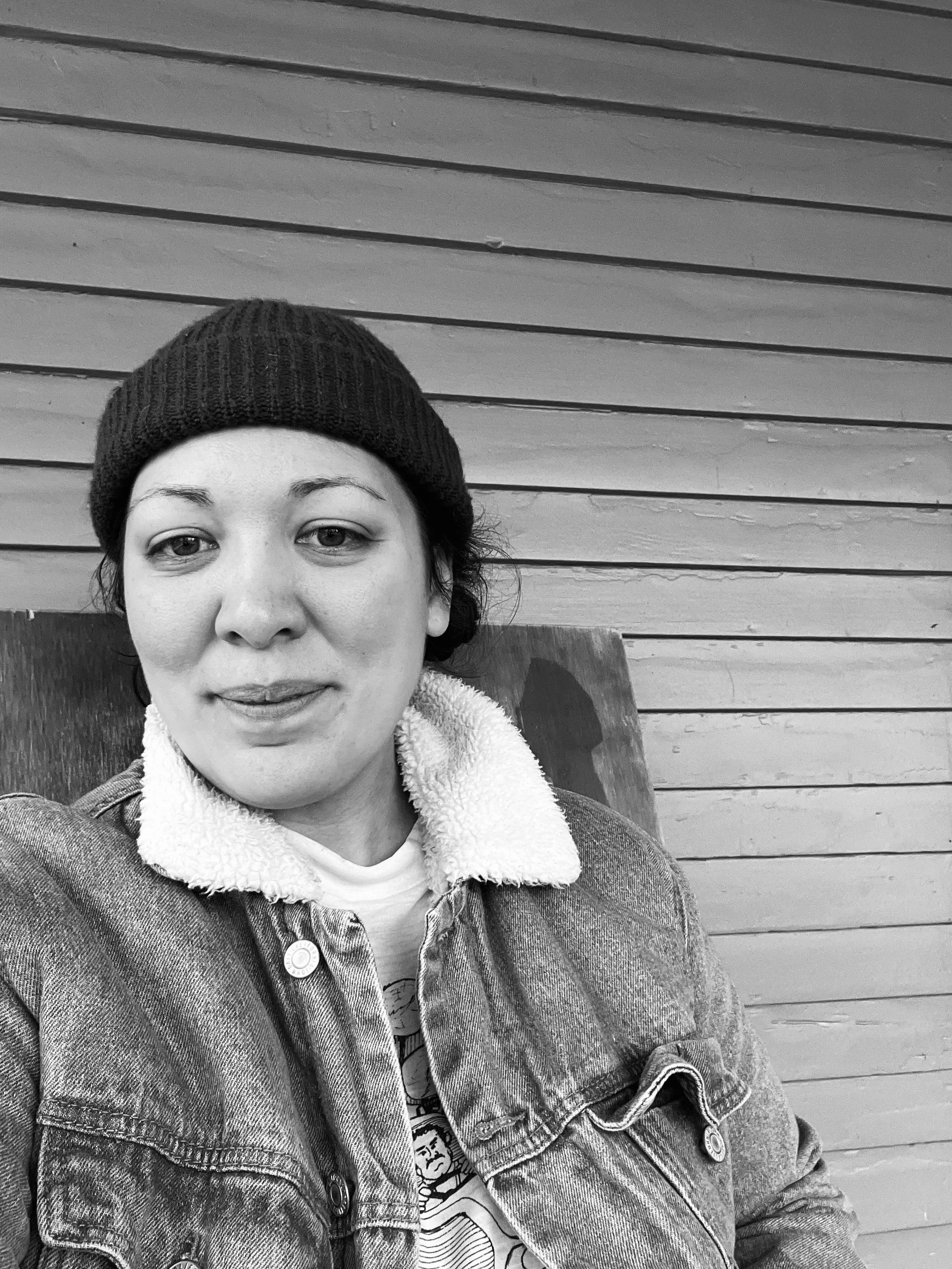 A young woman with short dark hair wearing a knit beanie, denim jacket with a fleece collar, and a graphic t-shirt, sitting against a wooden wall.