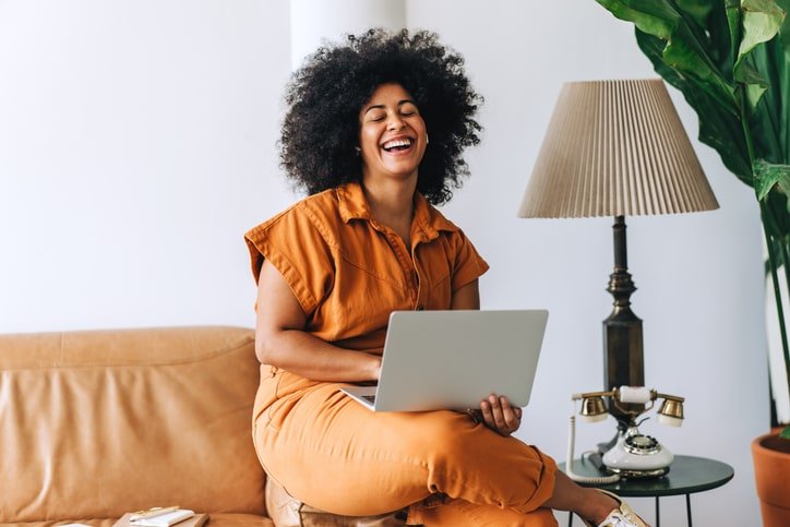 Black woman sitting on the arm of a couch laughing while using a laptop, representing online therapy for Black women in Chicago.