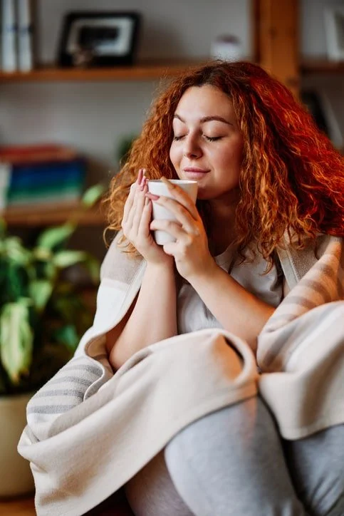 A woman wrapped in a blanket holding a warm mug at a kitchen table in soft morning light, symbolizing emotional exhaustion and burnout in January.