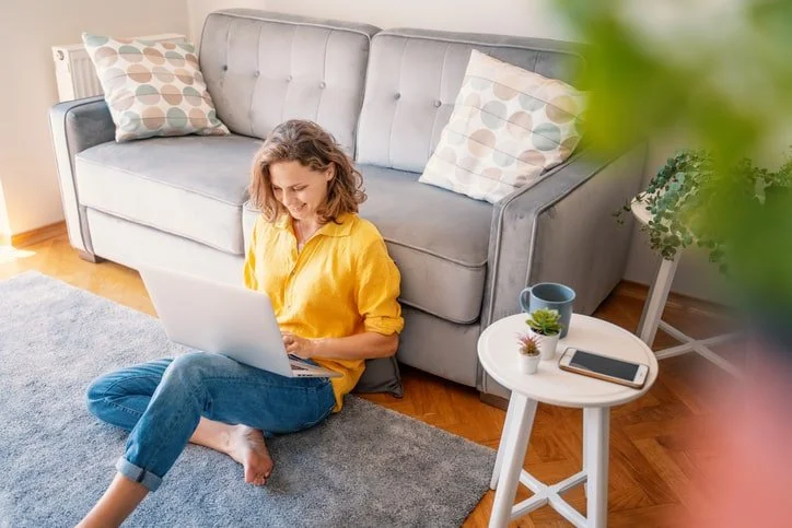 Woman sitting on the floor with a laptop in her lap and a couch behind her during an online therapy session, representing virtual mental health support in Chicago and Illinois.