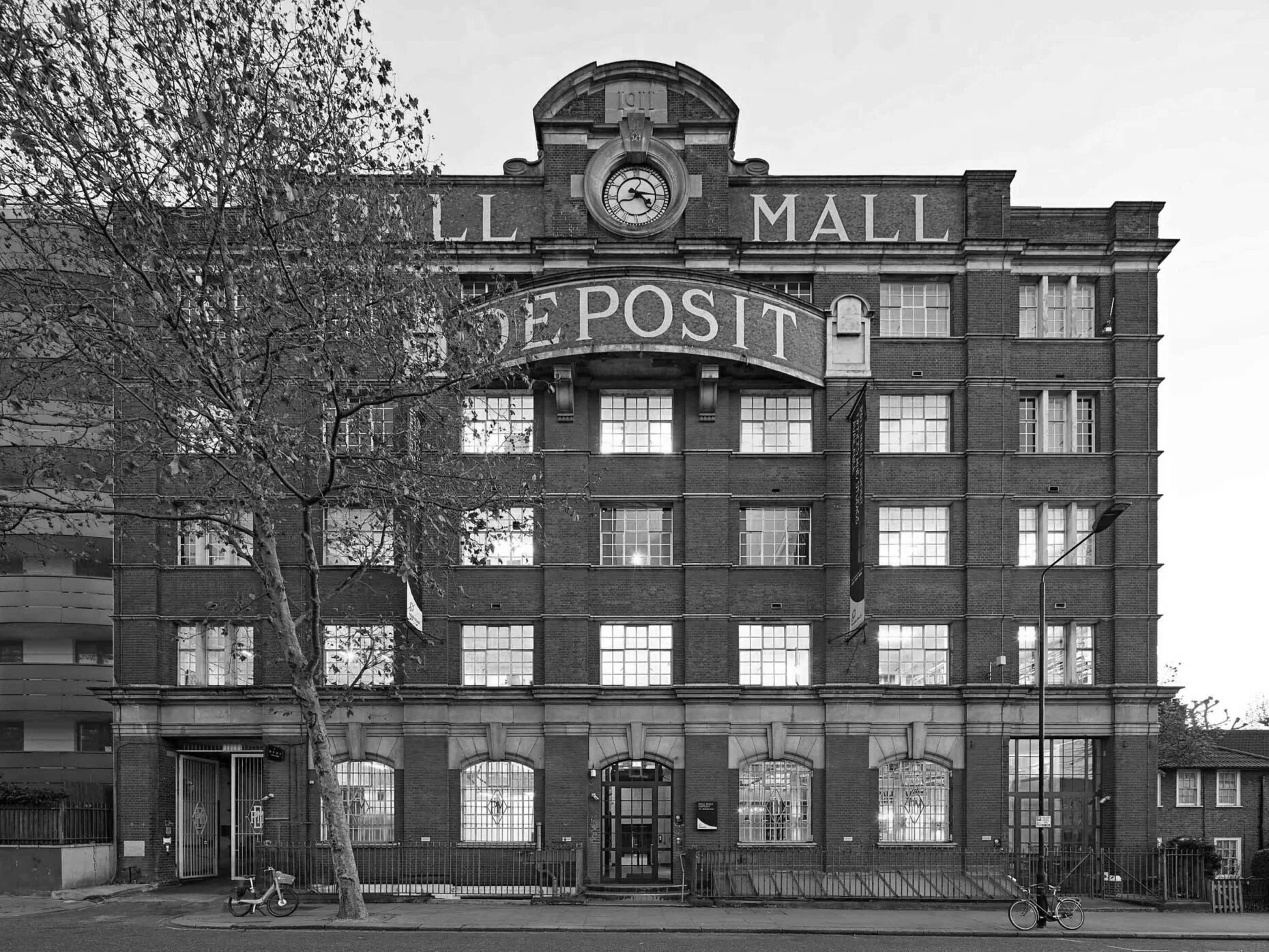 A black and white photo of a multi-story brick building labeled 'L. Mall' with a clock at the top and large windows. The building has an inscription that reads 'Depot' above the entrance and is surrounded by a small fence, with trees and bicycles in front.
