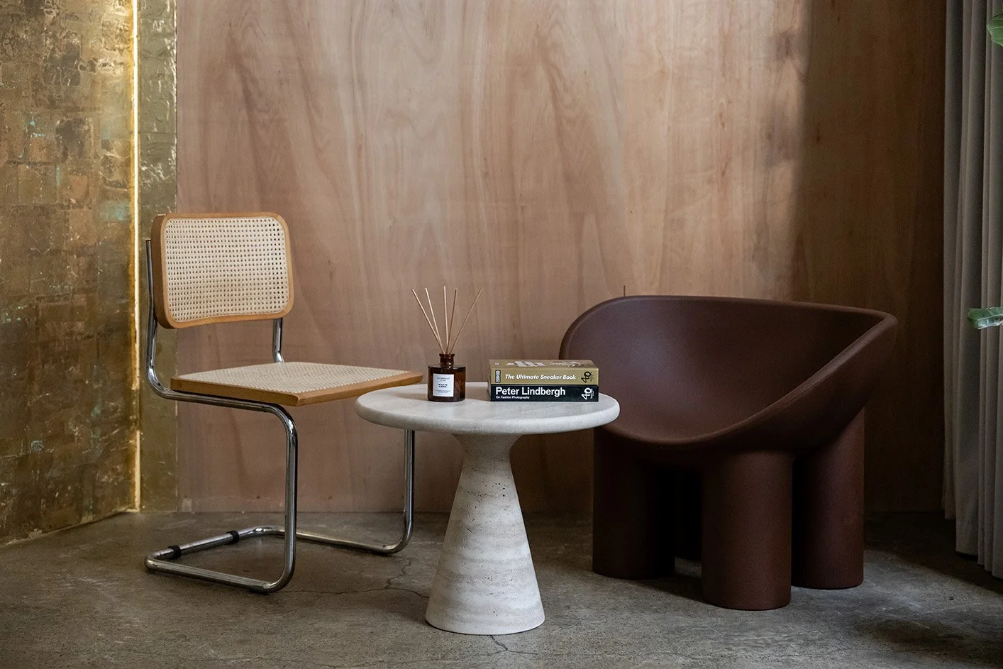 Minimalist interior with a beige chair, a marble-top side table, and a dark brown modern armchair against a pink textured wall, with books and reed diffuser on the table.