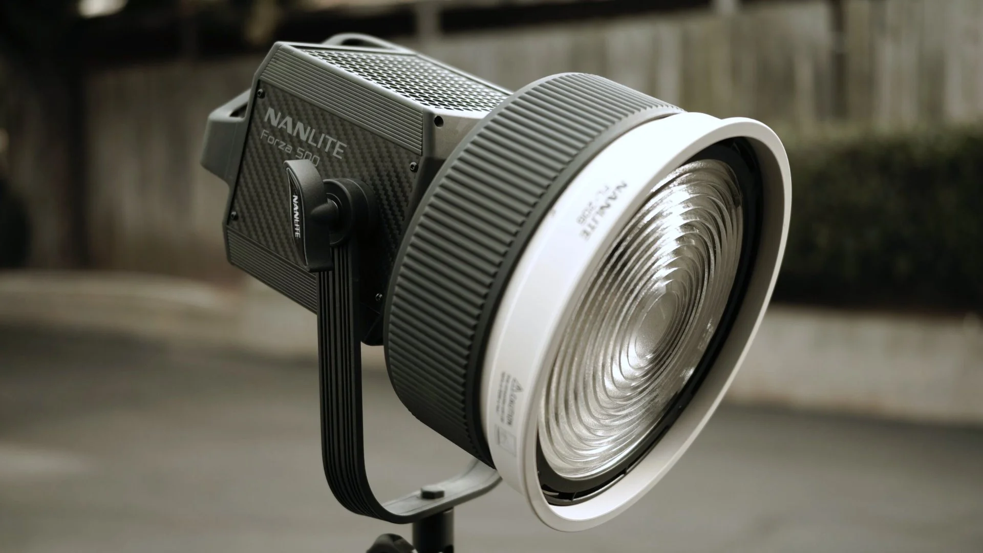 A professional stage or studio spotlight with a carbon fiber-like housing, labeled 'NANLITE Forza 500', mounted on a black stand outdoors, with a concrete wall and some greenery in the background.