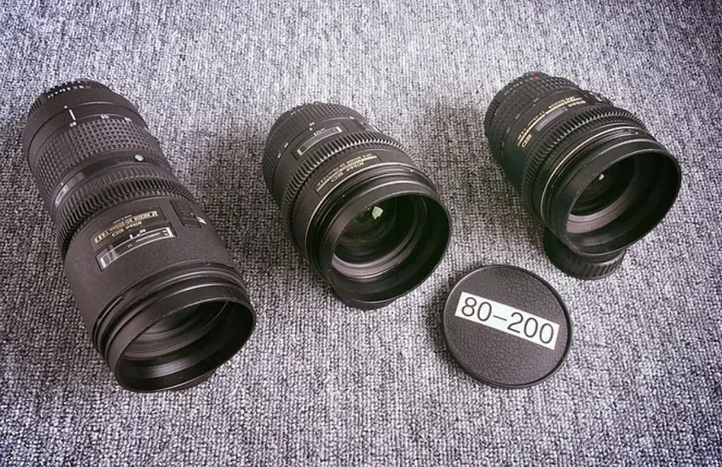 Three camera telephoto lenses placed on a textured gray surface, with a round lens cap marked "80-200" beside them.