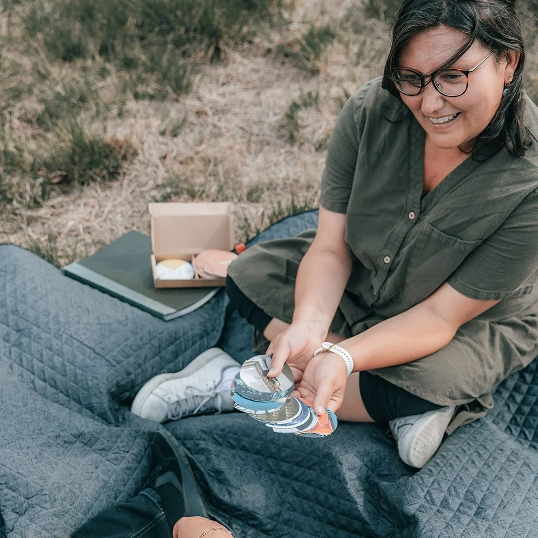 Leen Forment zit op een picknickdeken in de buitenlucht, houdt een set coachingkaarten vast en glimlacht, met een doos met accessoires en een boek naast haar.