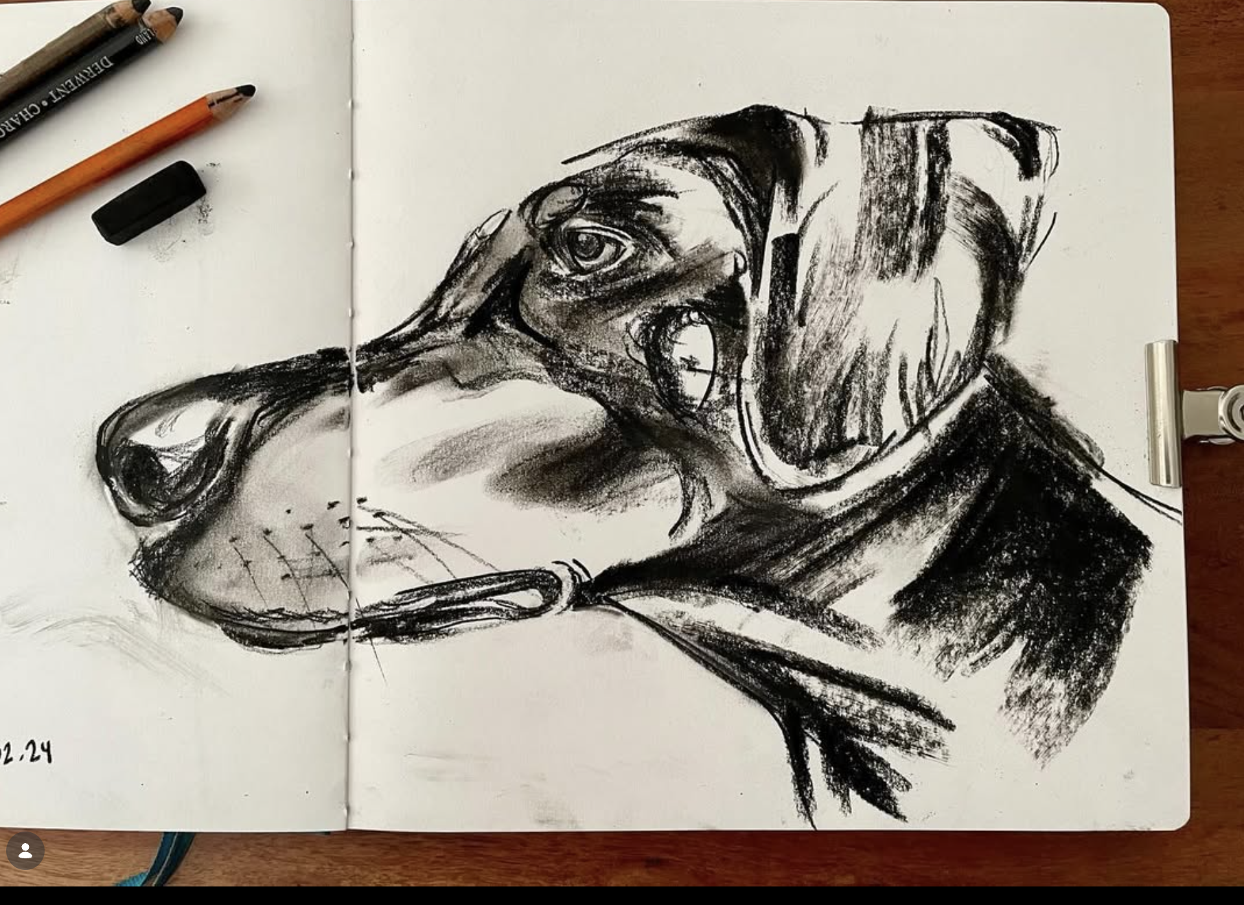Black charcoal sketch of a dog with its mouth open, showing teeth, on an open white sketchbook. Two pencils and an eraser are on the left side, and a clip on the right side of the sketchbook.