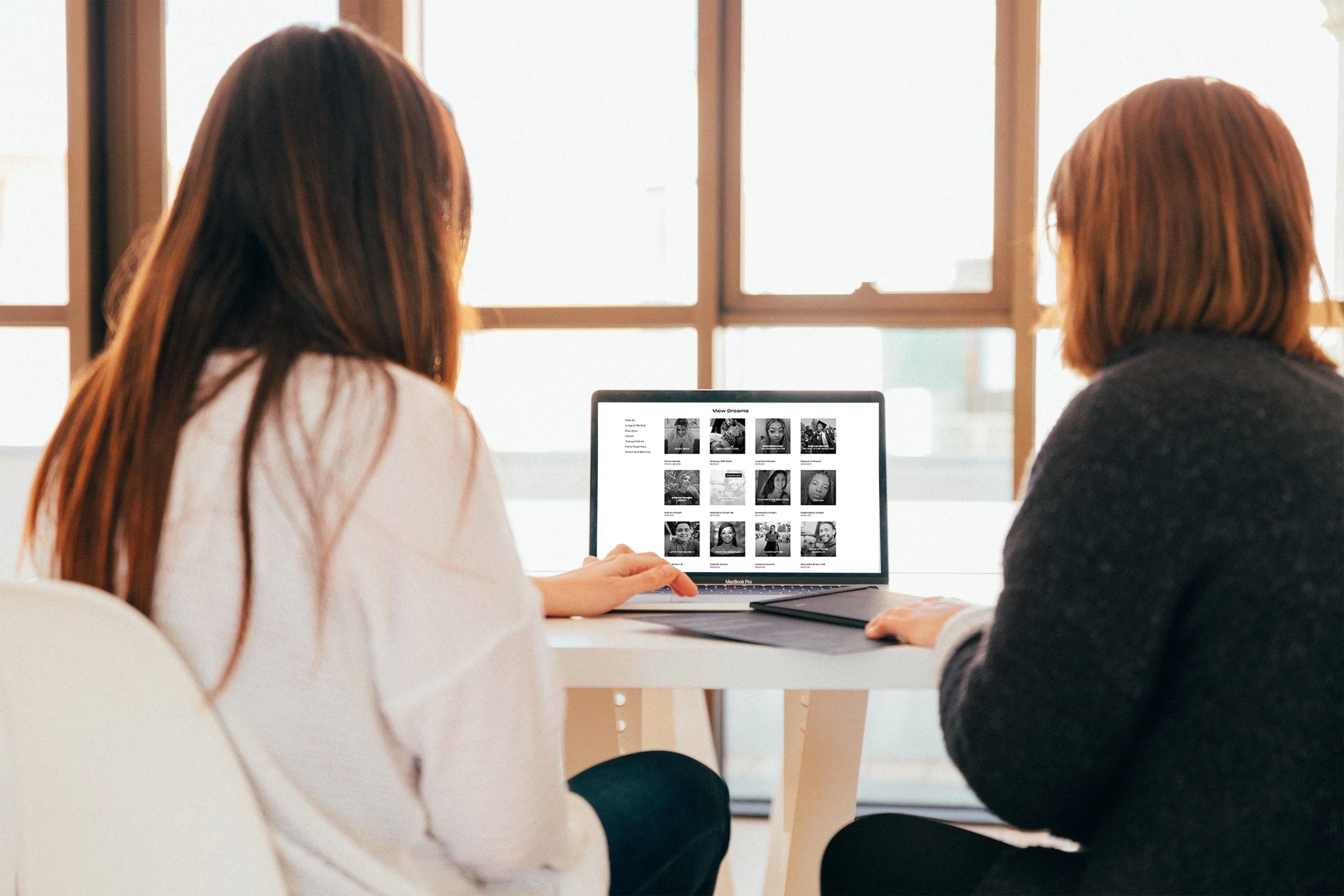 Two women sit at a white table, looking at a laptop displaying a grid of black-and-white profile pictures on a website or app, in front of large windows with a city view.