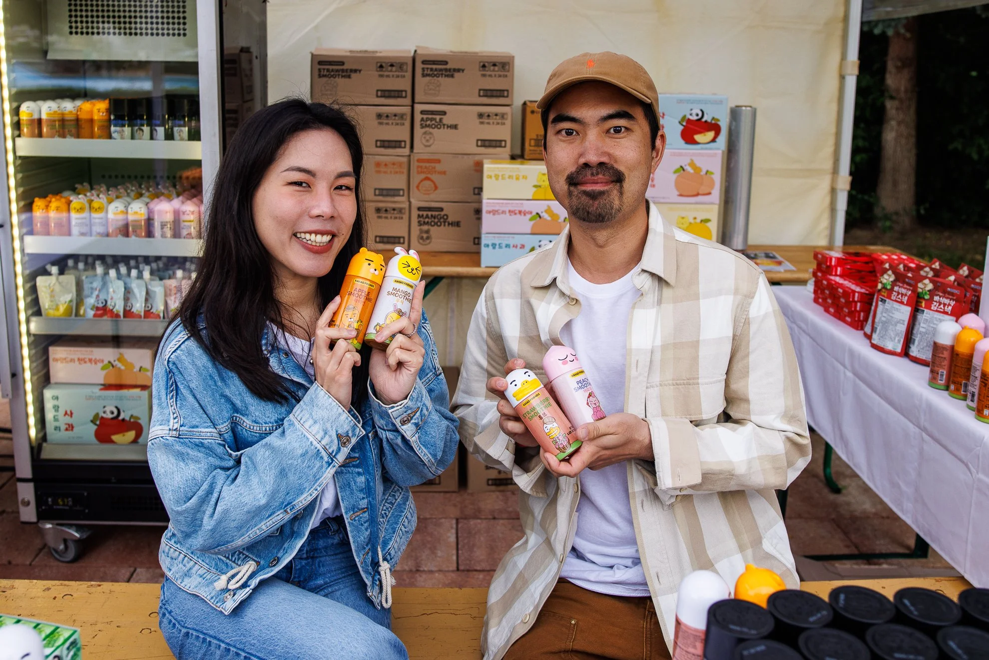 Zwei Menschen, eine Frau und ein Mann, stehen an einem Stand und präsentieren aufgelöste Flaschen mit niedlichem Tier-Design, umgeben von Produkten wie Fruchtshakes in Kartons hinter ihnen.