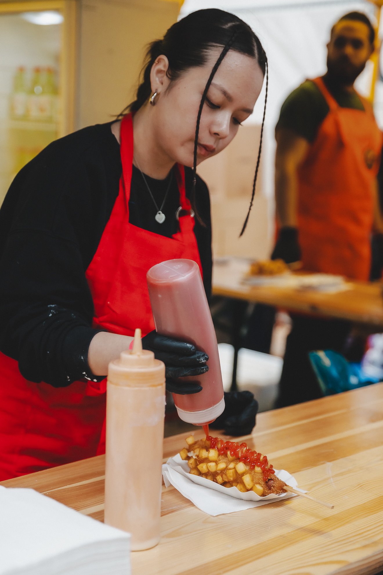 Eine Frau mit dunkler Haarfarbe, die eine rote Schürze trägt, bereitet Pommes Frites mit Sauce zu. Im Hintergrund ist ein Mann mit orangener Schürze zu sehen, der ebenfalls bei der Zubereitung von Speisen hilft.