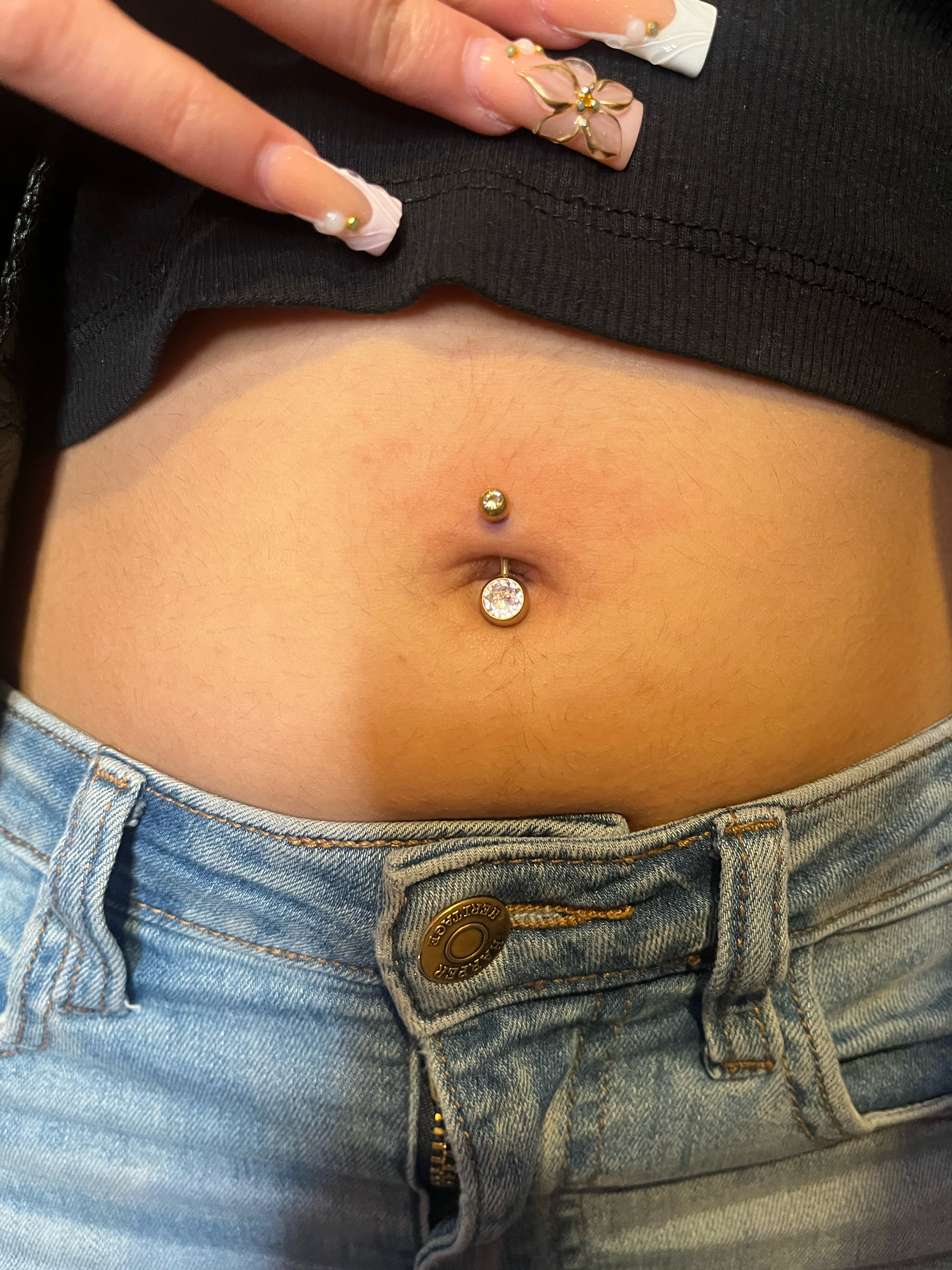 Close-up of a person's midsection showing a navel piercing with a large, clear gemstone and a smaller, yellow gemstone, with denim jeans and black clothing visible.