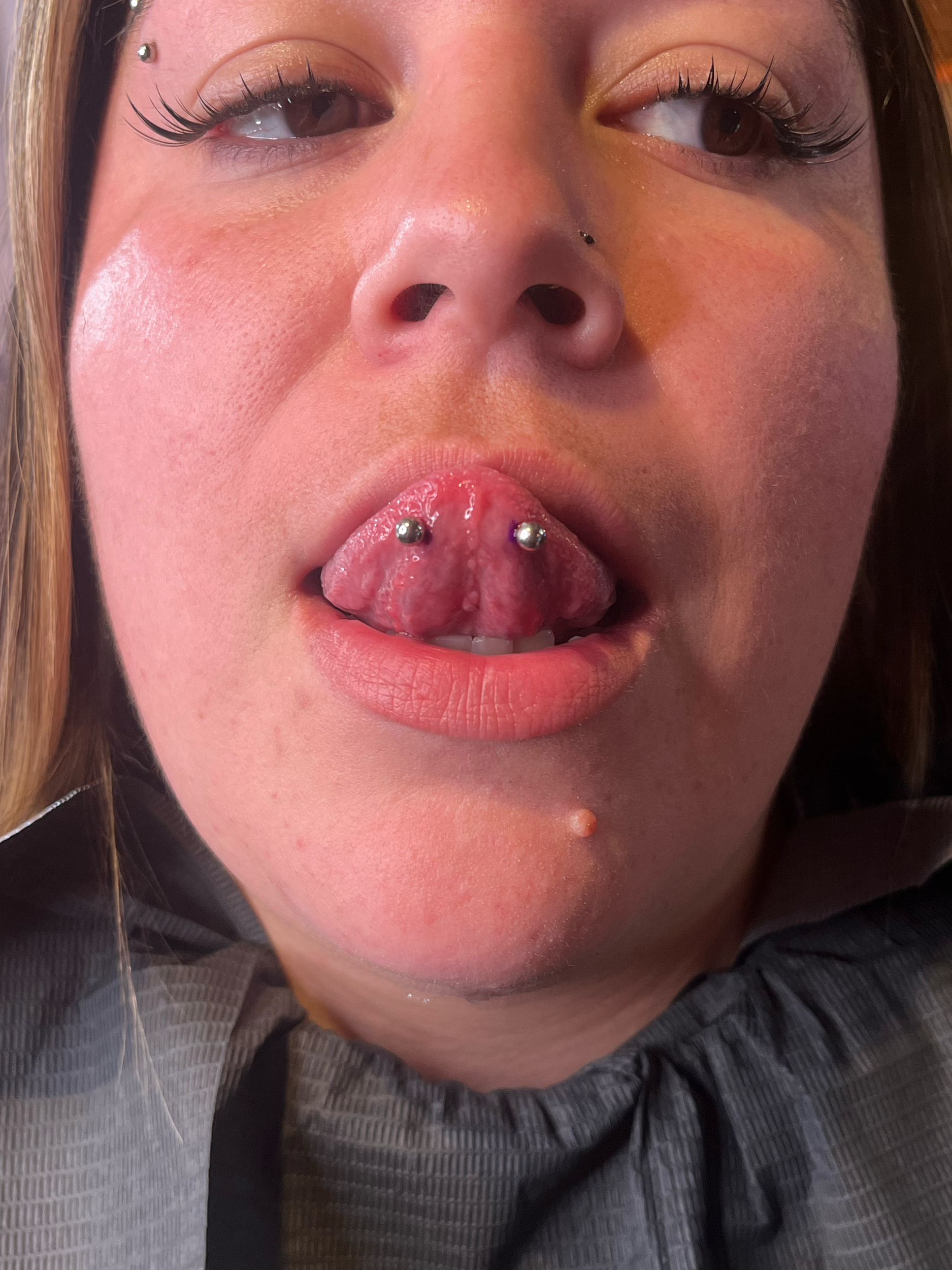 A woman with piercings and makeup lying down, her mouth open, showing her tongue with two piercings, and her chin and neck visible.
