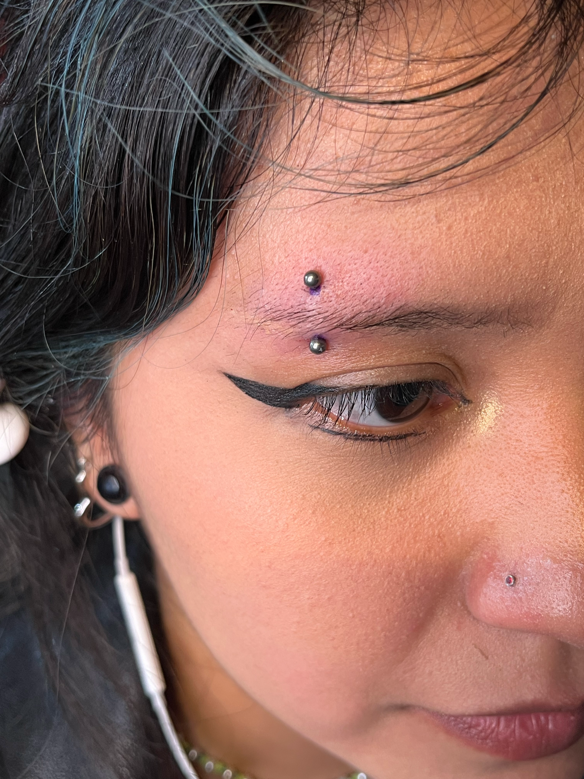 Close-up of a person's face with black hair, multiple facial piercings including a nostril and eyebrow piercing, and eye makeup with black eyeliner.