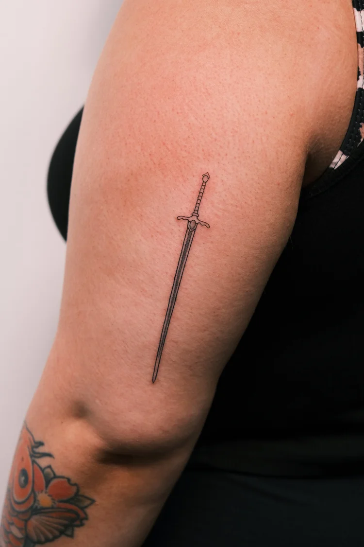 Fine line sword tattoo on an upper arm by Eli