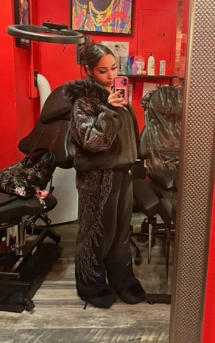 A young woman with dark hair wearing large white headphones, a black crop top, and blue jeans standing in a subway station.