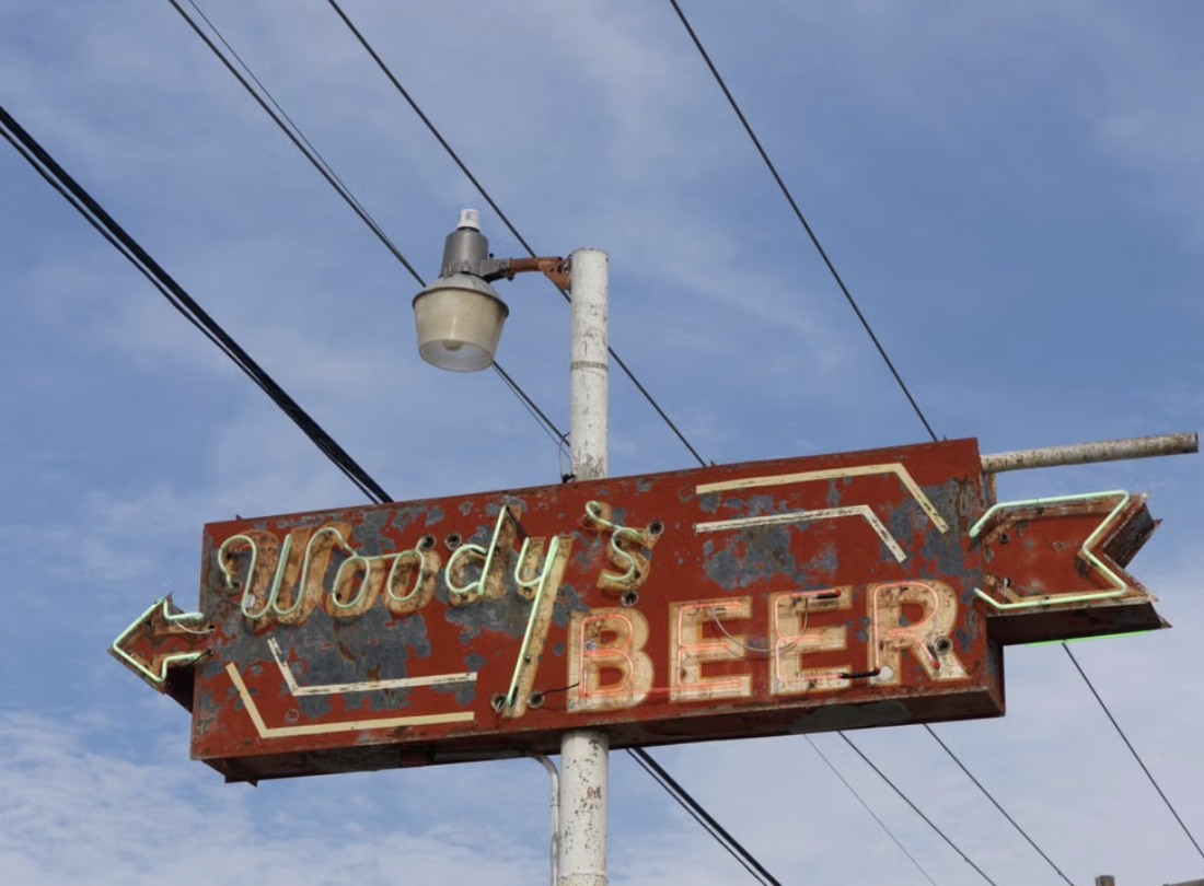 A vintage Americana-style sign for Woody's Beer, with a red background and signs of rust and age.