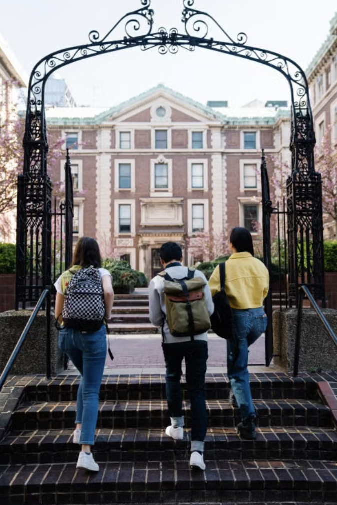 A group of three students with backpacks going up steps across an ornate archway, heading towards a university building in the background.