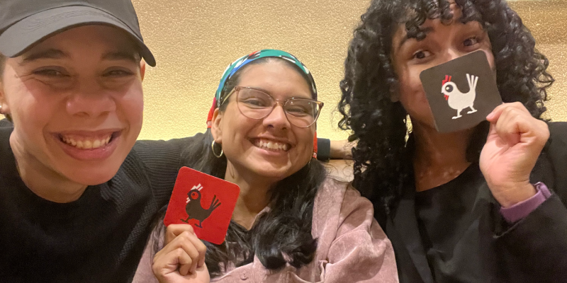 Layshi Curbelo, Karien Diaz y Yes Rosario sonriendo frente a la cámara en un restaurante. Dos sostienen pequeñas tarjetas ilustradas mientras posan juntas en un ambiente casual y alegre.