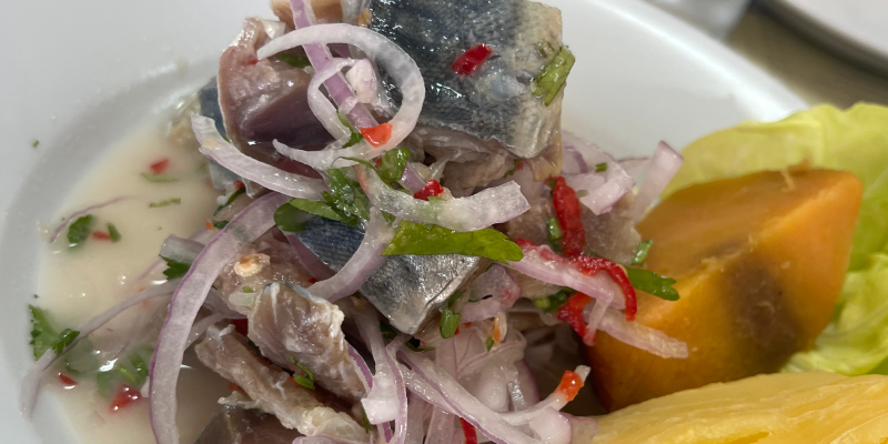 Plato de ceviche con pescado marinado, cebolla morada, ajíes y maíz, servido en un plato blanco con una rodaja de camote al lado.