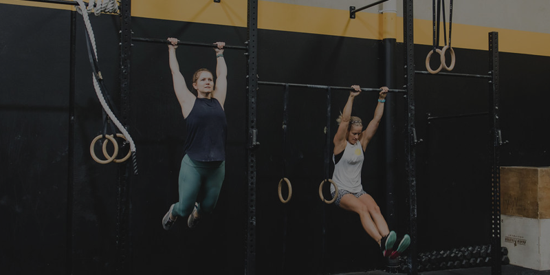 Two women hanging from gymnastic rings on a black and yellow gym wall.