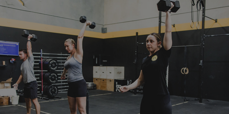 Three women lifting dumbbells overhead during a workout in a gym with black walls and fitness equipment.