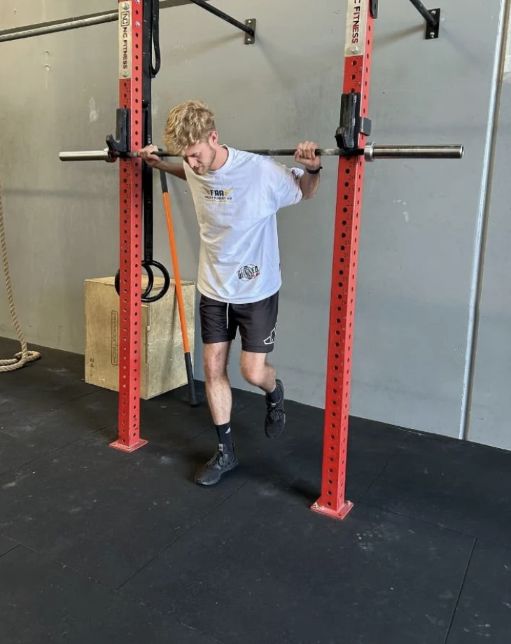 A young man performing a squat with a barbell at a gym, standing on one leg while holding the barbell across his shoulders.
