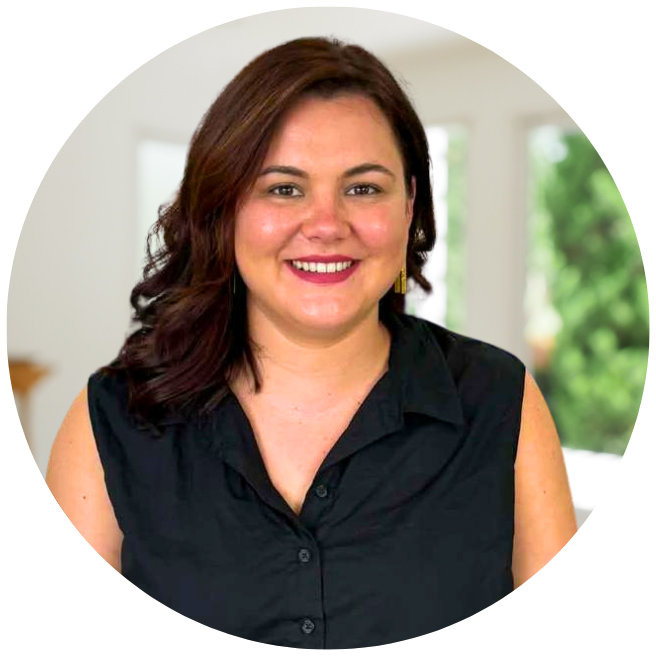 A woman with shoulder-length dark brown hair wearing a sleeveless black button-up shirt and gold earrings, smiling inside a bright room with large windows and greenery outside.