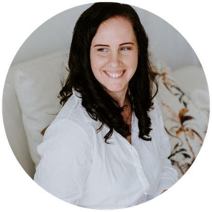 A woman with dark, curly hair smiling and looking to her left, sitting on a bed with white bedding and patterned pillows.