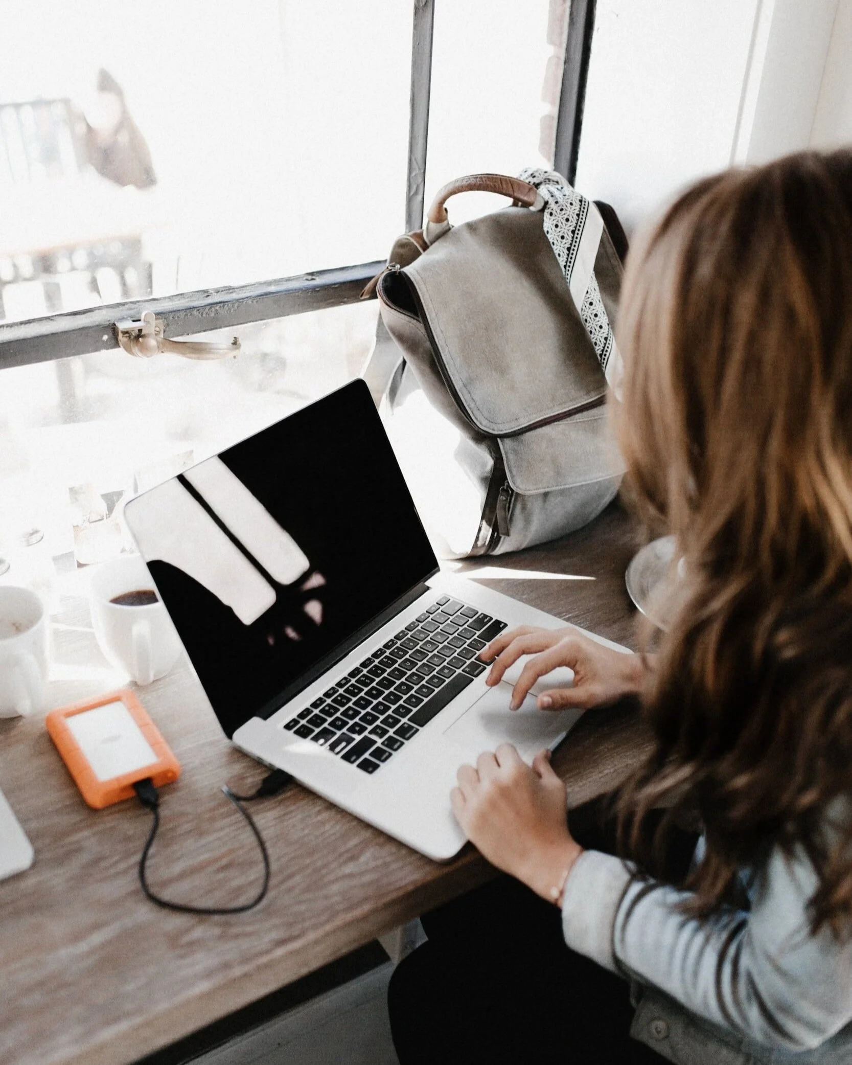 An eCommerce shop owner is checking her Pinterest marketing results on a laptop at a wooden desk with coffee cups, a portable charger, and electronic devices, sitting near a window.