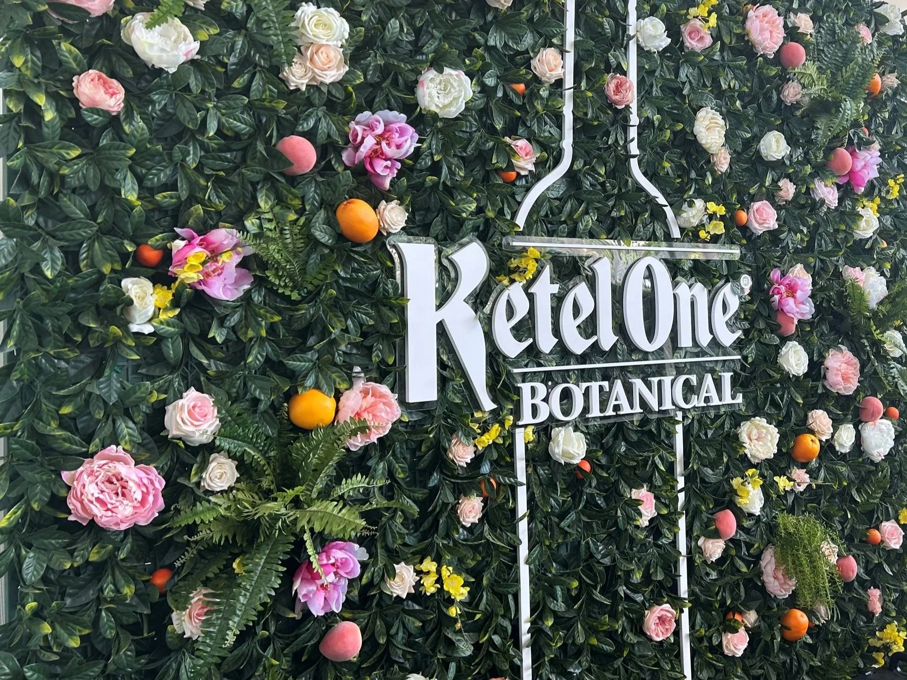A decorative wall of green foliage decorated with pink and white flowers and small orange fruits, with a white sign reading 'KetelOne Botanical' in front of it. corporate activation with this flower wall. 