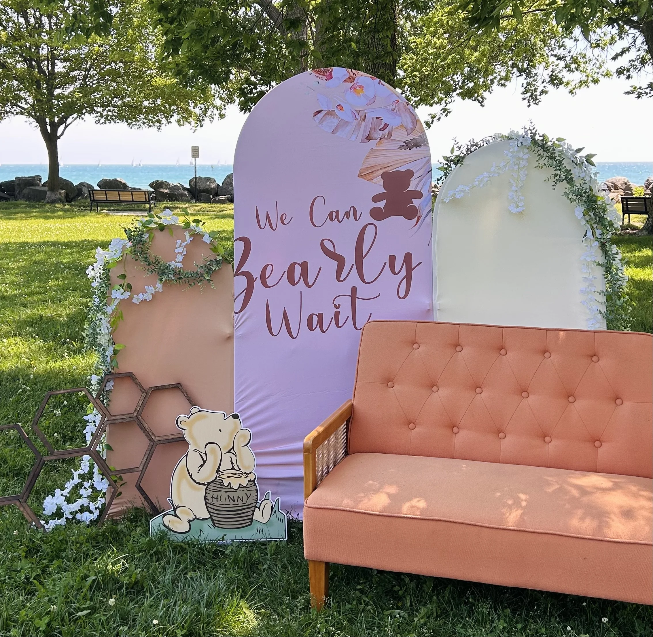 3 arches with fabric and a bright orange couch for the soon to be parents opening gifts at their baby shower. decorated with greenery, florals bears. a beautiful set up for a baby shower. 