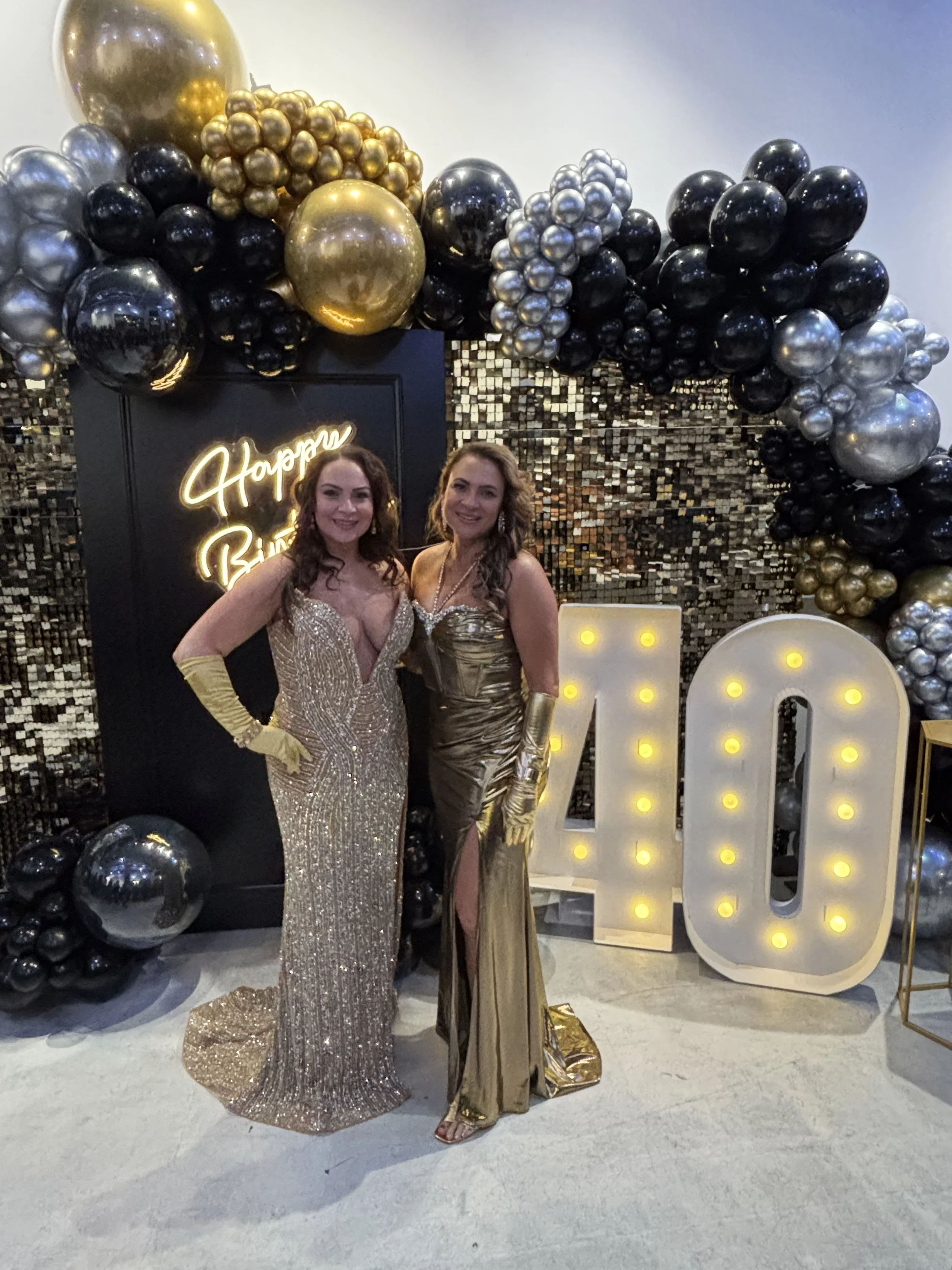birthday girls in front of their backdrop. 40th birthday backdrop. twins taking a photo in front of their sequence wall for their shared birthday party. milestone birthday celebration. 
