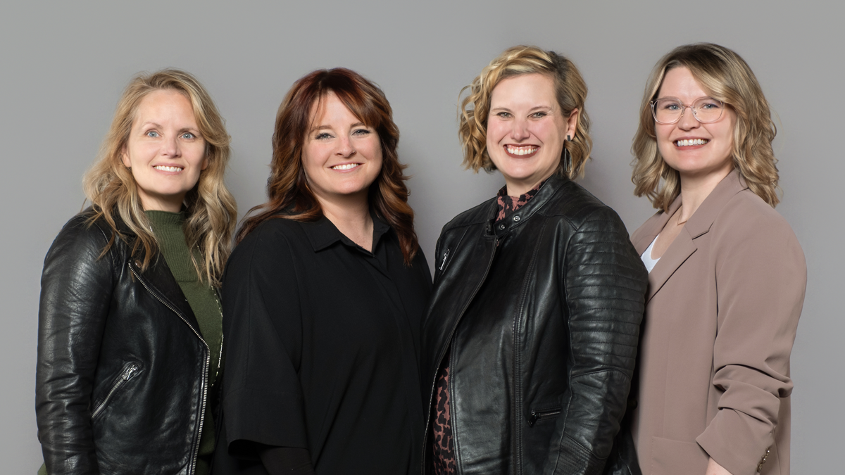 Four Women Owners of encompas furniture dealership in Kansas City, Omaha, and Wichita. Four white women of various ages wearing black and neutral colors, posing in front of gray background and smiling.