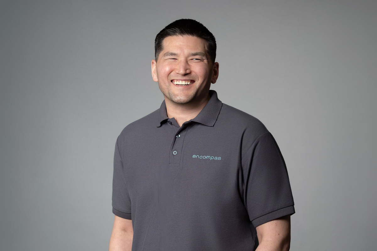 Travis Nishikawa, promoted at encompas Installation Company eIC from Installer to Installation Manager in 2026. Asian man with dark short hair, grinning and wearing a gray encompas branded polo shirt.