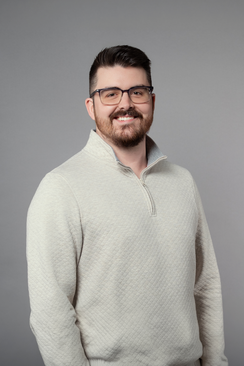 Austin Evans, promoted at encompas from Account Manager to Project Lead for Architectural Interiors in 2026. White man with glasses and dark brown trimmed beard standing relaxed and smiling, wearing a white quarter zip sweater.