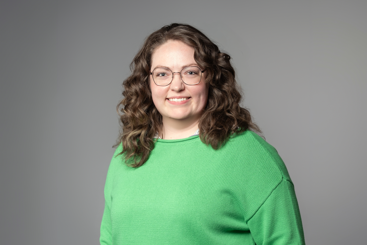 Carole Ollivierre, promoted at encompas from Marketing Coordinator to Communications and Brand Director in 2026. White woman with dark brown curly hair standing relaxed and smiling and wearing a bright green sweater.